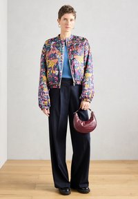 Veste bomber imprimée colorée avec une fermeture éclair à l'avant, pantalon long rayé et petit sac à main bordeaux à texture crocodile.