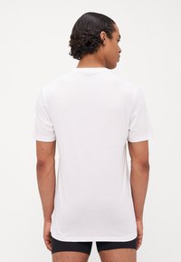 Weißes Baumwoll-T-Shirt mit kurzen Ärmeln, rundem Ausschnitt und glatter Textur, von hinten präsentiert, getragen mit schwarzen Shorts.