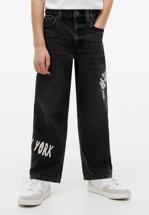 Άτομο που φοράει μαύρα τζιν παντελόνια με φαρδιά ποδιά με λευκό κείμενο "New York" και άσπρα αθλητικά παπούτσια, στέκεται μπροστά σε απλό φόντο.