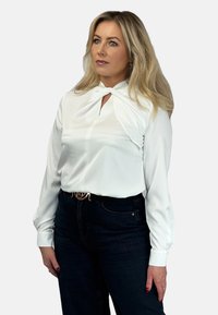 Biała bluzka z długim rękawem i detalem wiązania przy szyi, gładka faktura, w połączeniu z ciemnoniebieskimi dżinsami i czarnym paskiem z błyszczącą klamrą.