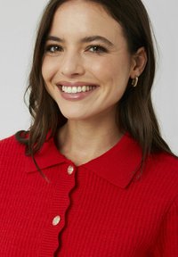 Cardigan en maille rouge texturée avec un col festonné, une fermeture à boutons dorés et des détails de design tissés.