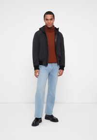 Veste à capuche noire avec une finition lisse, portée sur un pull marron côtelé, assortie à un jean bleu clair et des chaussures noires. Fond clair.