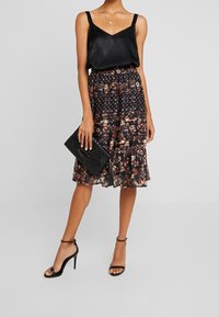 Camisole in satin nero, gonna in tessuto floreale a pois con orlo a ruches, abbinata a scarpe con tacchi a strappi neri e una pochette con texture.