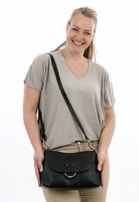 Svart läder crossbody-väska med texturerad yta, böjd magnetisk flap, och detaljer i guldton, hålls av en person mot en vit bakgrund.