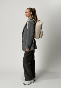 Blazer gris sur une chemise claire, associé à un pantalon sombre. Un sac à dos beige comprend plusieurs compartiments et une texture lisse. Baskets blanches.