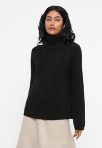 Črn rebrast pleten pulover s turtleneck ovratnikom in dolgimi rokavi, s sproščenim krojem in teksturirano tkanino, v kombinaciji s svetlo bež krilom.