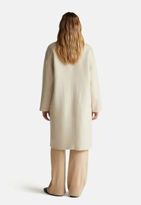 Lungo cappotto color crema realizzato in tessuto morbido, con spalle rilassate e una semplice cucitura sul retro, abbinato a pantaloni beige a gamba larga.