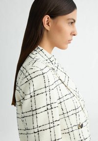 Blazer blanc avec motif écossais noir, tissu texturé, col cranté et accent de bouton noir unique, présenté en vue de profil latéral.