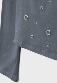 Ciemnoszara tkanina z rozrzuconymi srebrnymi metalowymi oczkami o różnych rozmiarach, pokazana z bliska z naciskiem na dolny brzeg i krawędź rękawa.