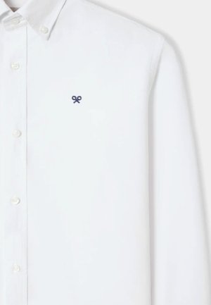 Camisa blanca de manga larga con botones y un pequeño logo azul marino en forma de lazo bordado en el pecho izquierdo.