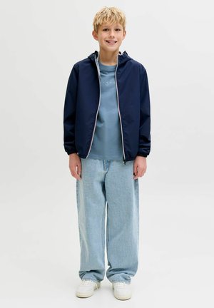 Blond chlapec sa usmieva, nosí tmavomodrú bundu s kapucňou cez svetlomodré tričko, voľné svetlozafarbené rifle a biele tenisky, stojí na jednofarebnom pozadí.