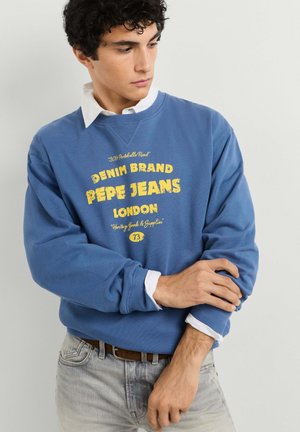 Joven ajustándose la manga de una sudadera azul con texto amarillo "Pepe Jeans London", lleva una camisa blanca con cuello y vaqueros gris claro.