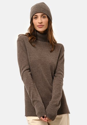 Donna che indossa un berretto di maglia marrone e un maglione a collo alto abbinato, con le mani giunte davanti, su uno sfondo chiaro e semplice.