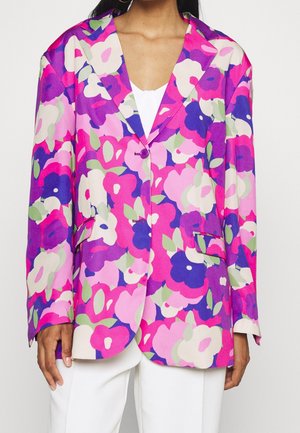 Woman wearing a bright floral blazer with pink, purple, white, and green flowers over a white top and white pants.