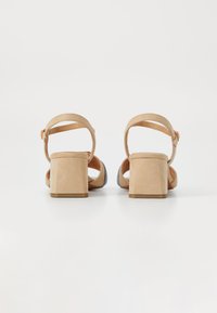 Sandalias de tacón cuadrado de ante beige con correas en el tobillo y backs redondeados. Superficie texturizada con un diseño minimalista y moderno.