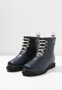 Botas de tobillo de goma azul oscuro con herrajes plateados, con una lengüeta alta y cordones, suela de goma negra y una cinta en la parte posterior para facilitar el uso.