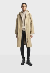 Kvinde står iført en beige hættefrakke, lysbeige lynlås-sweater, mørke leggings og sorte ankelstøvler på en ensfarvet lys baggrund.