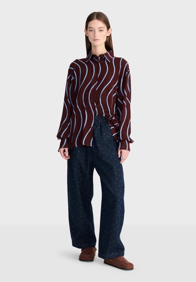 Jeune femme debout portant une chemise bordeaux à rayures ondulées bleues, un pantalon ample foncé et des chaussures marron sans lacets, sur un fond uni.