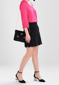 Donna che indossa una blusa rosa brillante, una gonna nera con bordi a scallop, una borsa nera con decorazioni a forma di ape e tacchi neri con blocchi a pois bianchi.