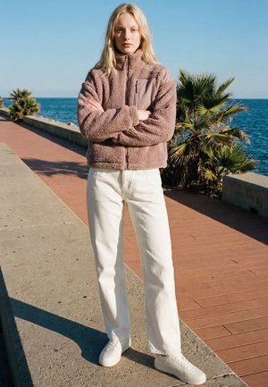 Fiatal nő összefont karokkal, barna polár dzsekit és fehér nadrágot viselve, tengerparti sétány mellett áll, pálmafákkal.