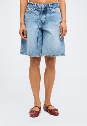 Stredne dlhé svetlomodré denimové šortky so zviazaným opaskom v páse, nosené s červenými plochými topánkami na osobe na jednofarebnom pozadí.