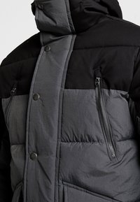 Giacca a vento imbottita con design a blocchi di colore nero e grigio, caratterizzata da collo alto, bottoni a scatto e tasche sul petto con cerniera. La texture appare imbottita.