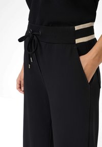 Schwarze Hose mit einem Kordelzugbund mit metallenen Spitzen, Seitentaschen und einem strukturierten Bund mit cremefarbenen und schwarzen Streifen.