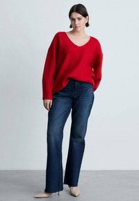 Maglione rosso a maglia con scollo a V, abbinato a jeans scuri a vita alta con orlo sfilacciato. La modella indossa scarpe beige a punta.
