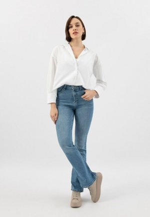Chemise blanche à manches longues avec boutons, jean taille haute bleu, et baskets beiges claires à lacets, présentés sur un fond uni.
