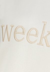 Witte stof met opvallende beige geborduurde tekst "week." Gladde textuur met een subtiele glans.