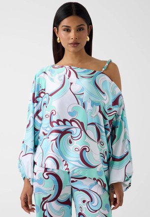 Femme portant un haut ample et un pantalon assortis avec des motifs abstraits tourbillonnants en bleu, sarcelle, blanc et marron, boucles d'oreilles en or et bretelle perlée.