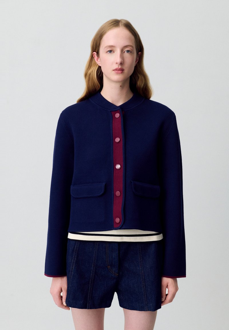 Giacca a coste blu navy con colletto rotondo, patta dei bottoni bordeaux e due tasche anteriori. Indossata sopra una maglietta a righe bianche e blu navy.