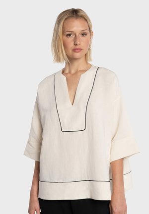Blouse en lin beige avec un col en V, des manches courtes, et un passepoil noir soulignant le col et l'ourlet, créant un contraste marqué.