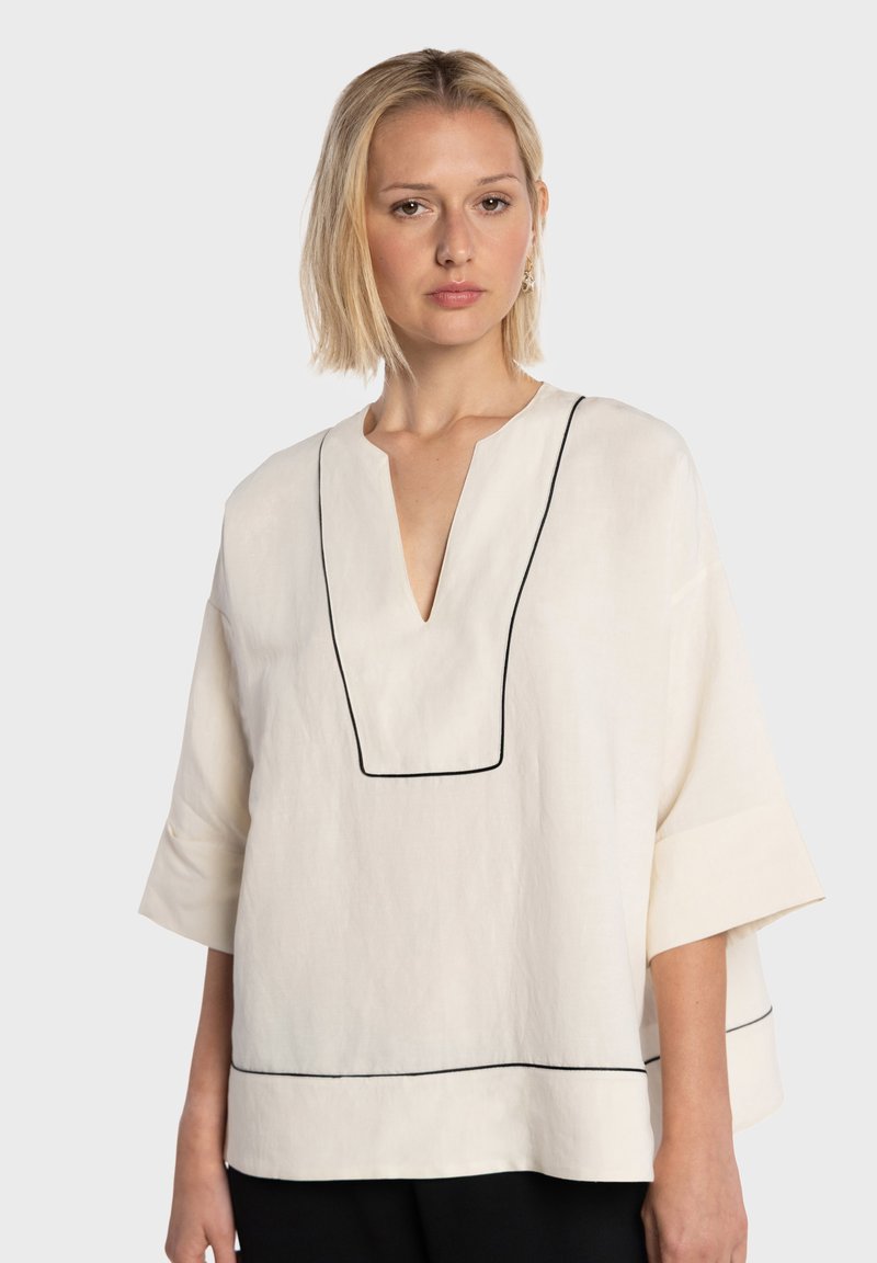 Blouse en lin beige avec un col en V, des manches courtes, et un passepoil noir soulignant le col et l'ourlet, créant un contraste marqué.