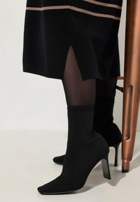 Bottines noires avec un design ressemblant à des chaussettes, bout pointu et talon aiguille haut, associées à un legging noir et une robe à rayures horizontales.