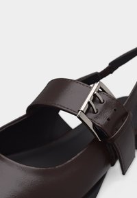 Sandale en cuir marron foncé avec une large lanière et une fermeture à boucle argentée, dotée d'un design à bride arrière et d'une finition lisse.