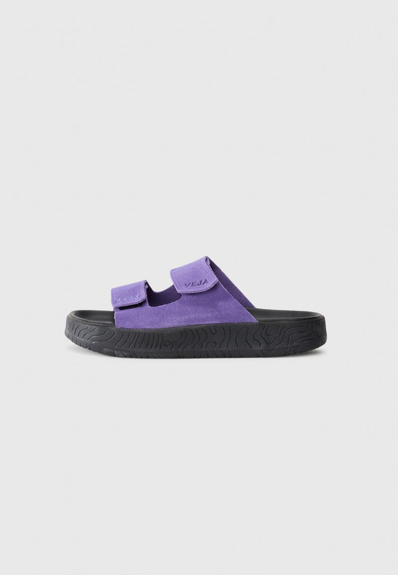 Lila mockaskinns slide-sandal med dubbla Velcro-remmar och svart strukturerad sula, visad i sidoprofil mot en enkel bakgrund.