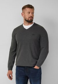 Grijze V-hals trui gemaakt van een zacht materiaal met lange mouwen, met een subtiel logo op de borst, gedragen over een wit T-shirt.