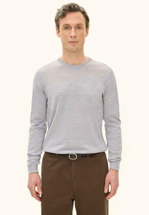 Mann steht, trägt ein hellgraues Langarmshirt und braune Hose mit schwarzem Gürtel, schaut nach vorne vor einfachem Hintergrund.