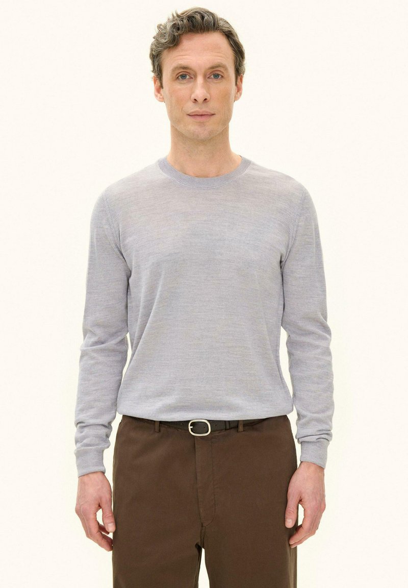 Mann steht, trägt ein hellgraues Langarmshirt und braune Hose mit schwarzem Gürtel, schaut nach vorne vor einfachem Hintergrund.