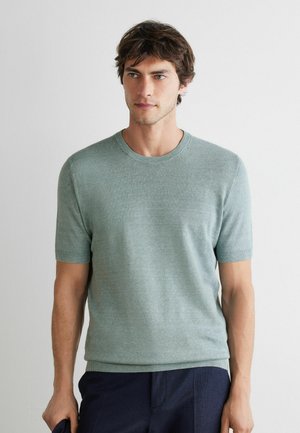 Junger Mann, der einen hellgrünen kurzärmeligen Strickpullover und dunkle Hosen trägt, steht vor einem schlichten hellen Hintergrund.