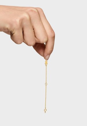 Ręka trzyma delikatny złoty łańcuszek z małą zawieszką w kształcie misia na jednolitym białym tle.