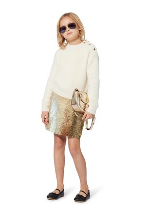 Maglione bianco, morbido e a maniche lunghe con bottoni dorati, abbinato a una mini gonna dorata e lucida. Scarpe nere e una borsa a tracolla in oro metallico.