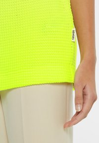 Neon-gelbes Stricktop mit einem texturierten Muster, das an der Seite ein Etikett mit der Aufschrift "SENSES" aufweist. Der Stoff wirkt leicht und atmungsaktiv.