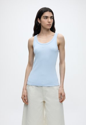 Γυναίκα με μακριά σκούρα μαλλιά φοράει ανοιχτό μπλε αμάνικο τοπ και άνετο λευκό παντελόνι, στέκεται μπροστά σε απλό λευκό φόντο.