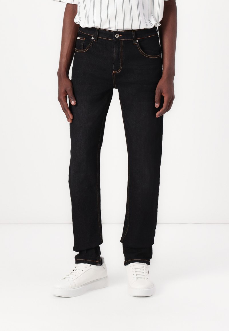 Schwarze Jeans aus Denim mit orangen Nähten, Five-Pocket-Design, gerade Passform, getragen mit weißen Sneakers und einem gestreiften Hemd.