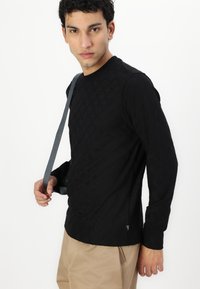 Black long-sleeve shirt with a textured pattern, round neckline, and a slim fit. Paired with beige trousers and a grey strap across the shoulder.