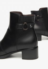 Zwarte leren enkelboots met lage blokhakken, zijdecoratieve riem en metalen ringdetail op een witte achtergrond.