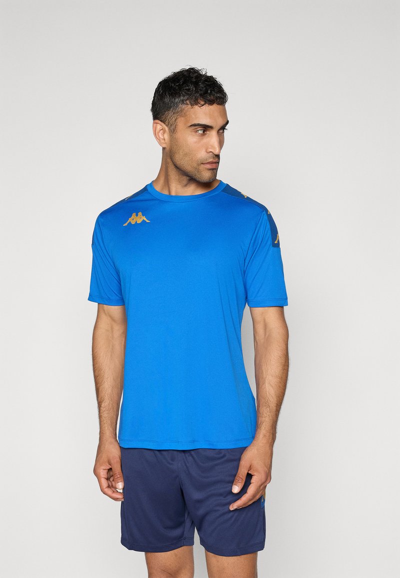 Mann iført en blå trenings-T-skjorte med gullfargede logoer på skuldrene og mørkeblå shorts, som ser mot venstre mot en ensfarget bakgrunn.