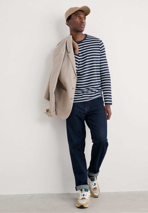 Camisa de rayas azul marino y blanca de manga larga, blazer beige colgado sobre el hombro, jeans azul oscuro y zapatillas multicolores con acentos.